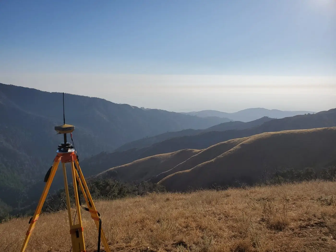 GNSS receiver on a tripod on a grassy ridge above the Big Sur backcountry, with the Pacific haze beyond