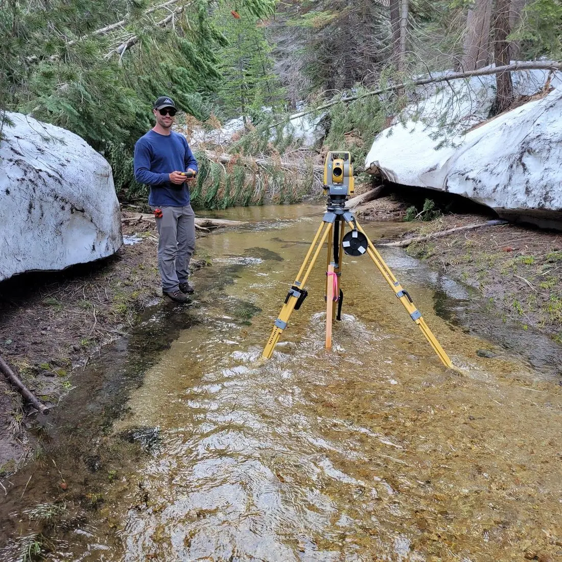 Surveyor standing in a shallow snowmelt creek beside a total station on a tripod