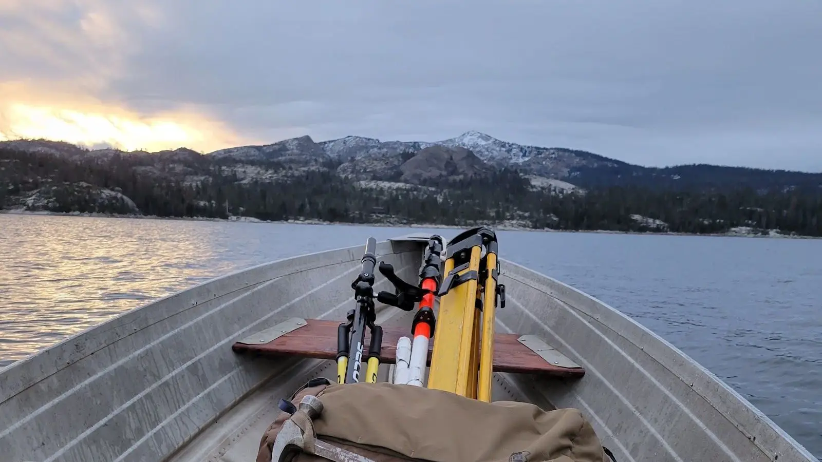 Survey tripods, rods and instrument cases loaded in an aluminum boat crossing a remote alpine lake toward snow-dusted peaks at dawn