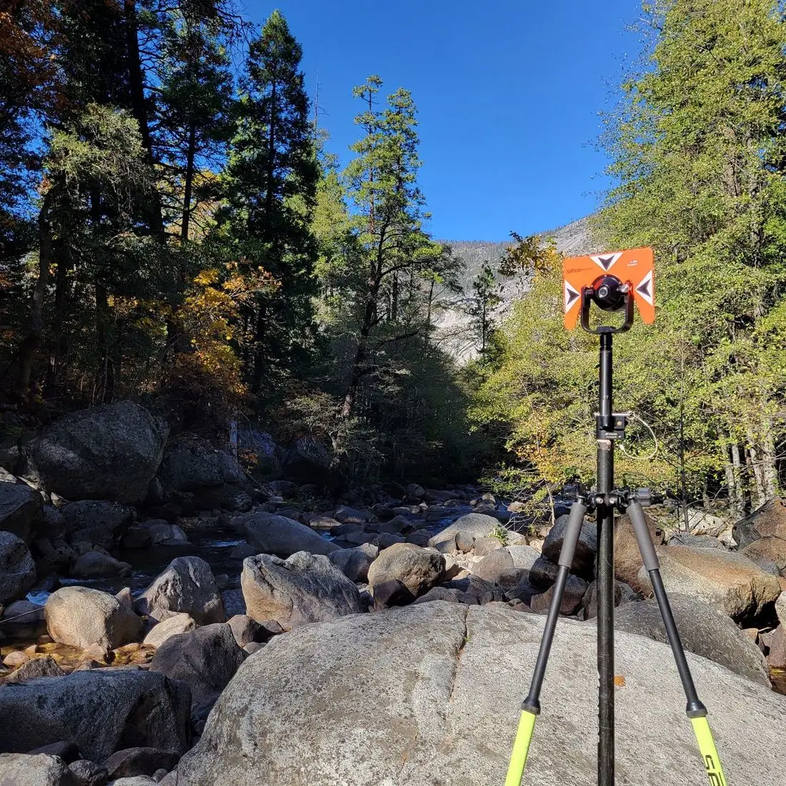 Orange survey prism on a tripod above a granite riverbed under pines
