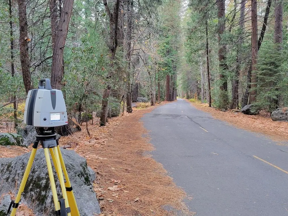Laser scanner on a yellow tripod beside a paved forest road covered in fallen pine needles