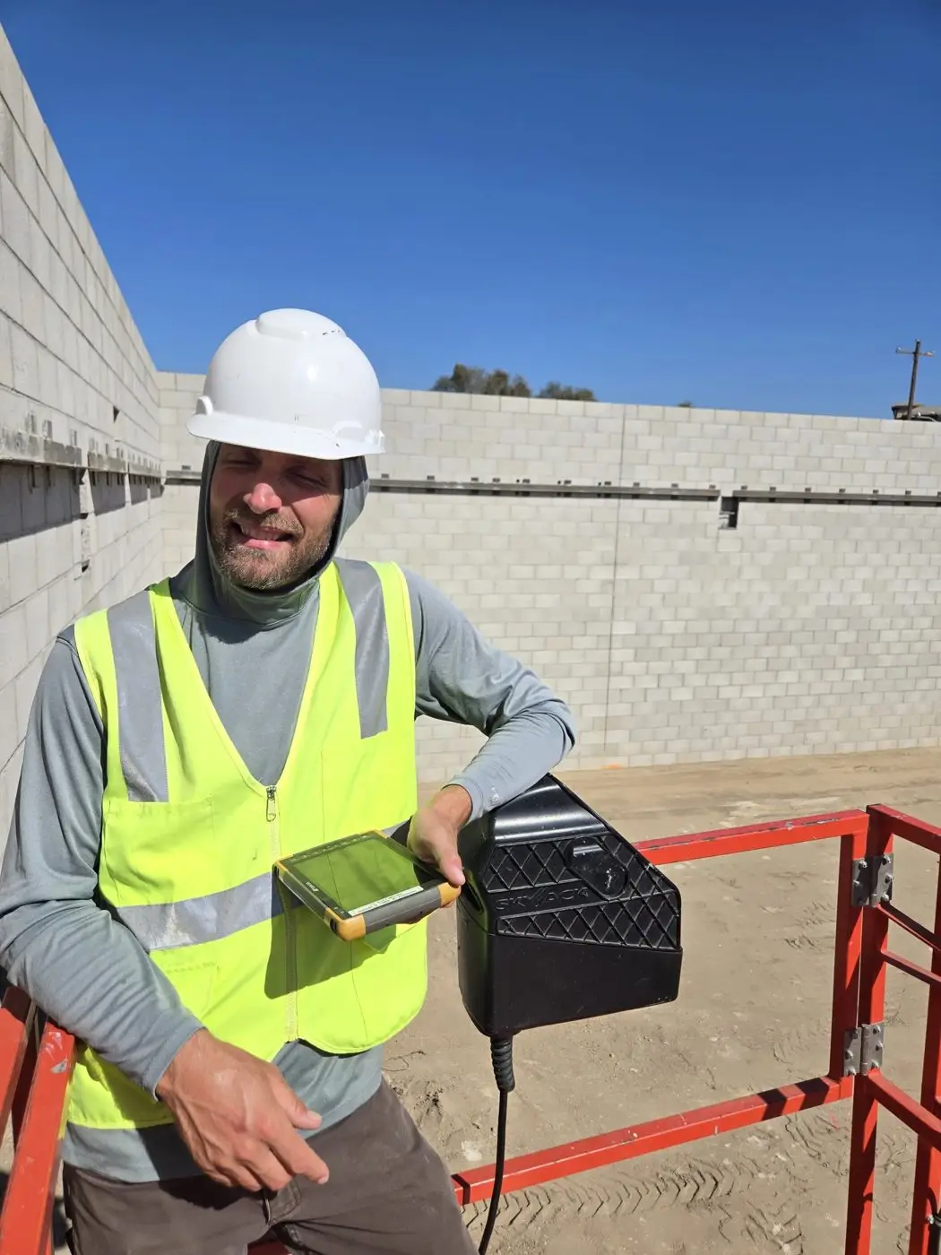 Licensed surveyor in a hard hat and safety vest on a lift with a data collector, beside a new block-wall building