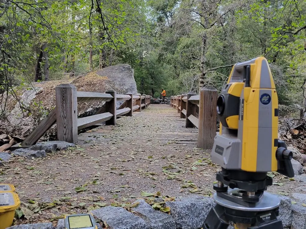 Robotic total station set up on a trail beside a wooden fence and granite boulders