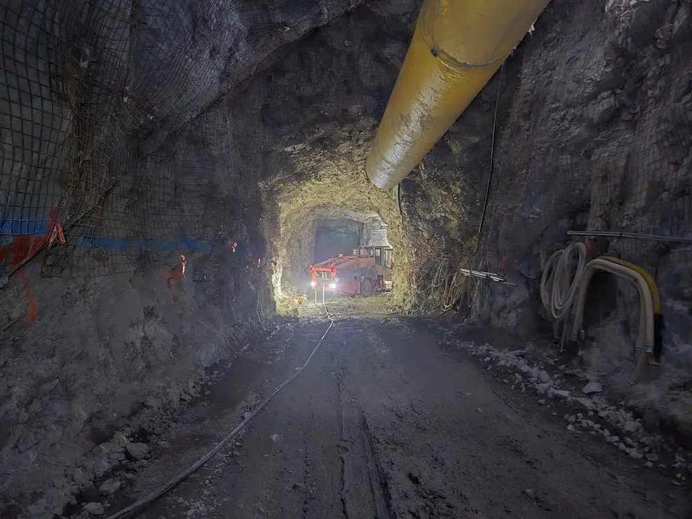 Underground decline tunnel with a drill jumbo working at the lit heading, survey paint marks on the rib and a ventilation duct overhead